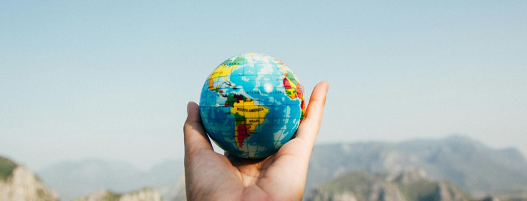 Hand hält einen kleinen Globus vor einer Berglandschaft und blauem Himmel